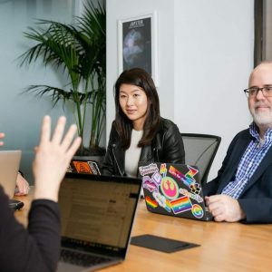 boardroom meeting where woman in black leather jacket and man in blue checkered shirt, suit jacket and glasses, pay attention to person talking with their hands wearing black long sleeve shirt